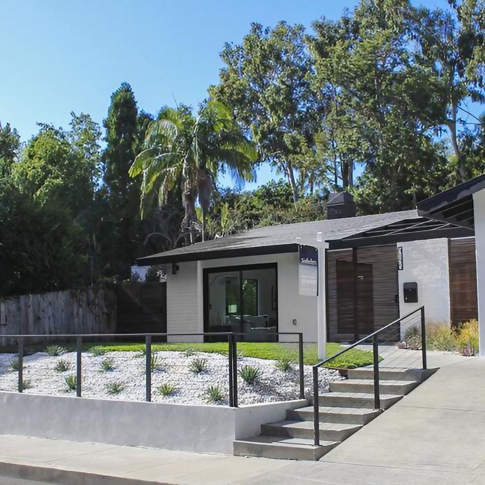 Detached house with flat roof, white facade and wood paneling, surrounded by palm trees and other trees, with stairs and railing leading to the entrance.